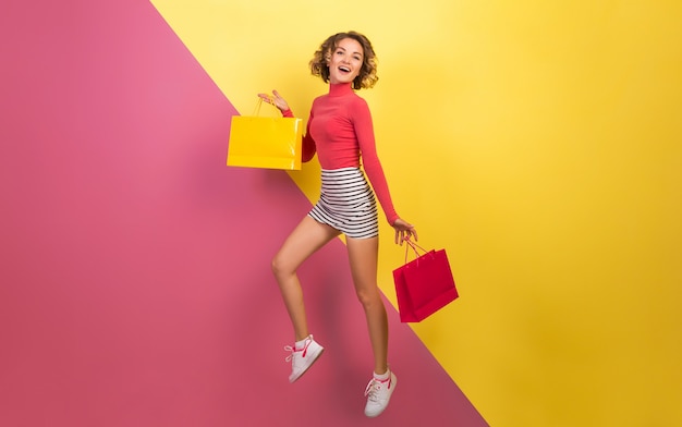 Gratis foto glimlachende aantrekkelijke vrouw in stijlvolle kleurrijke outfit springen met boodschappentassen