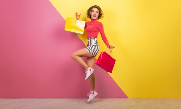 Glimlachende aantrekkelijke vrouw in stijlvolle kleurrijke outfit springen met boodschappentassen