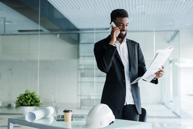 Glimlachende Afrikaanse Amerikaanse zakenman die op de telefoon met krant en kop van koffie in bureau spreekt