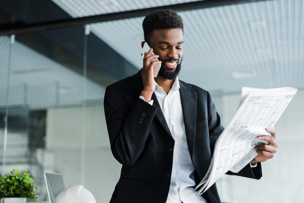 Glimlachende Afrikaanse Amerikaanse zakenman die op de telefoon met krant en kop van koffie in bureau spreekt