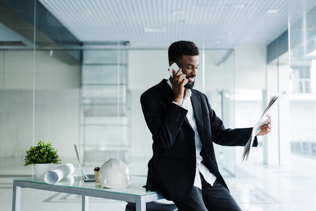 Glimlachende Afrikaanse Amerikaanse zakenman die op de telefoon met krant en kop van koffie in bureau spreekt