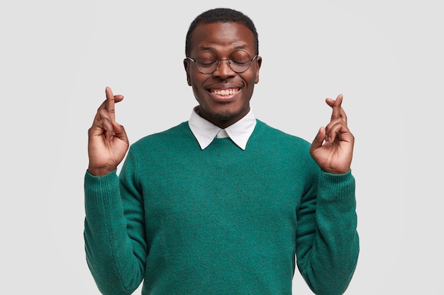 Glimlachende Afro-Amerikaanse man bidt voor de vervulling van zijn wens, kruist vingers, wenst veel succes