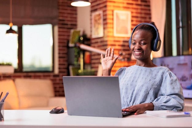 Glimlachende Afro-Amerikaanse vrouwelijke externe werknemer groet collega's op videocall in modern kantoor aan huis. Videoconferentievergadering met bedrijfsteam, zakenvrouw in koptelefoon op teleconferentie