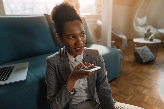 Glimlachende Afro-Amerikaanse zakenvrouw die over de luidspreker van de mobiele telefoon praat terwijl ze thuis werkt