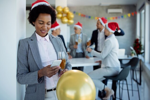 Glimlachende Afro-Amerikaanse zakenvrouw sms't op mobiele telefoon tijdens kerstfeest op kantoor