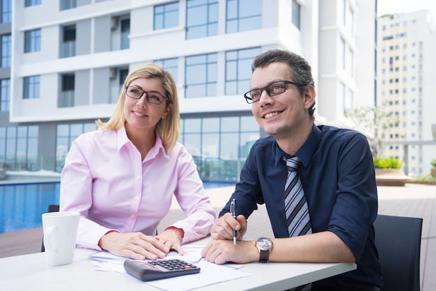 Glimlachende ambitieuze accountants die bij lijst met documenten en calculator zitten