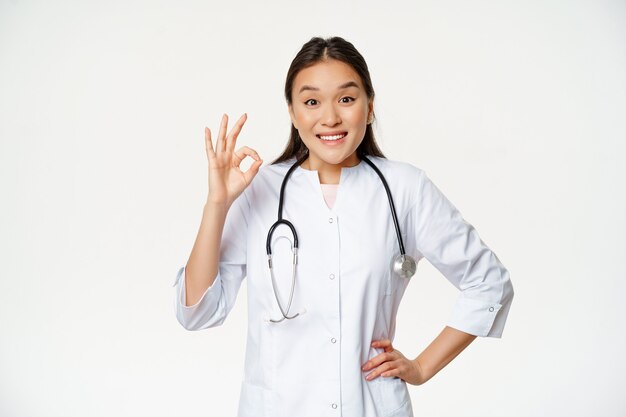 Glimlachende aziatische arts vertoont een goed teken, draagt een medisch gewaad. Vrouwelijke ziekenhuismedewerker in uniform beveelt iets aan, staande op een witte achtergrond.