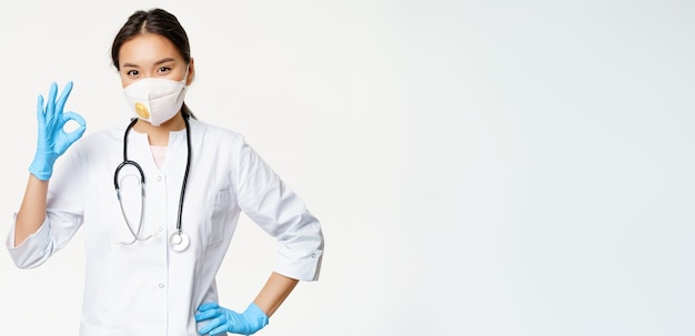 Glimlachende aziatische arts-vrouwelijke arts toont goed teken draagt ademhalingsapparaat van het vangen van covid-ziekte s