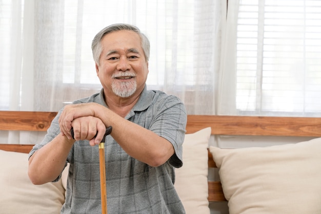 Glimlachende aziatische bejaarde man die gezond met pensioen gaat en een stok vasthoudt en ontspant terwijl hij thuis op de bank in de woonkamer zit