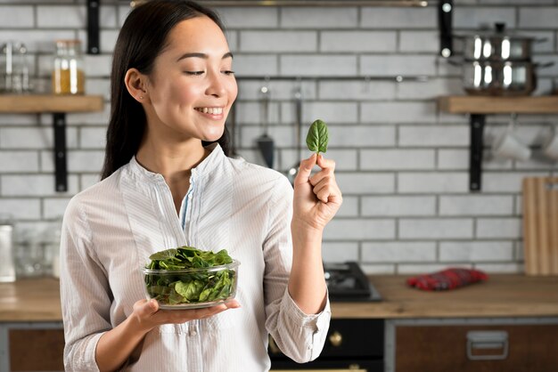 Glimlachende Aziatische vrouw die basilicumblad in keuken bekijkt