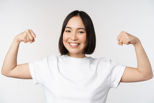 Glimlachende aziatische vrouw die buigende bicepsspieren toont, sterk wapengebaar dat zich in witte t-shirt op witte achtergrond bevindt