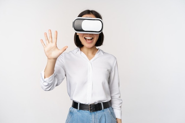 Glimlachende aziatische vrouw die naar je zwaait met een virtual reality-bril vr-headset die chat in meta-universum over een witte achtergrond