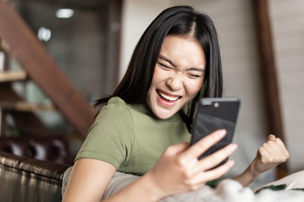 Glimlachende aziatische vrouw die tevreden kijkt, vuistpomp maakt en mobiele telefoon leest om thuis te zitten en ...