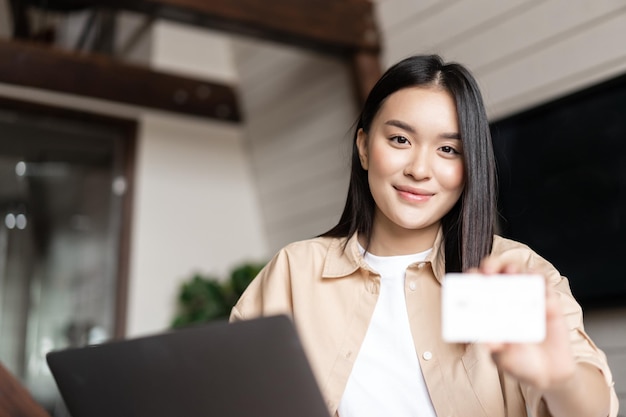 Glimlachende aziatische vrouw die thuis zit met computer die creditcard toont bij cameraconcept online sh...