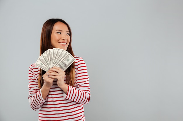 Glimlachende Aziatische vrouw in het geld van de sweaterholding en weg het kijken over grijze achtergrond