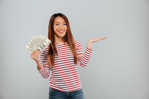 Glimlachende Aziatische vrouw in sweater die geld tonen en copyspace op het pond over grijze achtergrond houden