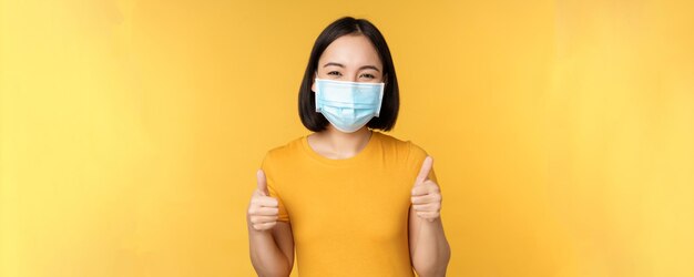 Glimlachende aziatische vrouw met een medisch gezichtsmasker met duimen omhoog, zoals en aanbevelen om over een gele achtergrond te staan