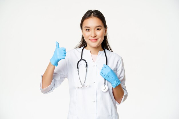 Glimlachende aziatische vrouwelijke arts toont duimen, draagt rubberen handschoenen en kliniekuniform, staat op een witte achtergrond