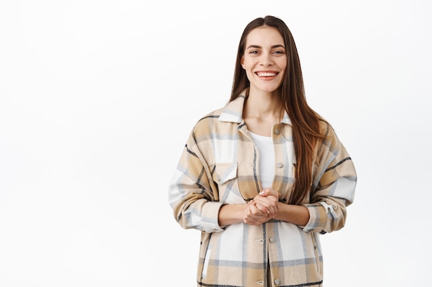 Glimlachende beleefde vrouw klaar om te helpen, wil helpen, handen bij elkaar houden over de borst en vriendelijk naar voren kijkend, toespraak houdend, tegen een witte muur staan