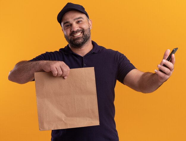 Glimlachende bezorger van middelbare leeftijd in uniform en pet met papieren voedselpakket met telefoon geïsoleerd op gele muur