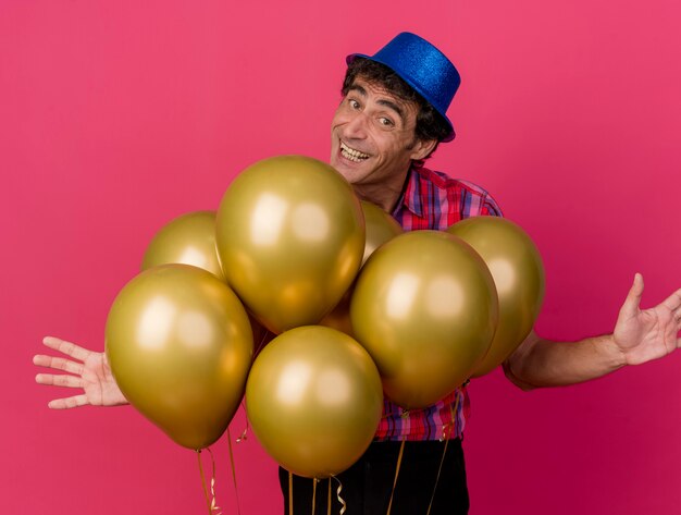 Glimlachende blanke feestmens van middelbare leeftijd met feestmuts achter ballonnen kijken naar camera met lege handen geïsoleerd op een karmozijnrode achtergrond