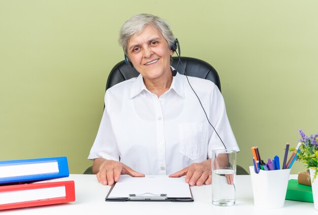 Glimlachende blanke vrouwelijke callcenter-operator op koptelefoon zittend aan bureau met kantoorhulpmiddelen met klembord geïsoleerd op groene muur