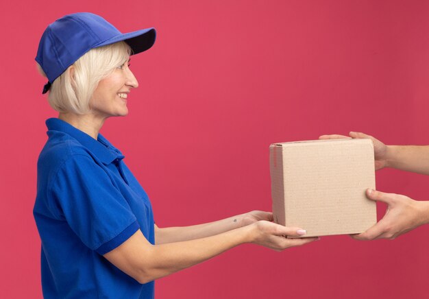 Glimlachende blonde bezorger van middelbare leeftijd in blauw uniform en pet die in profielweergave staat en kartonnen doos geeft aan klant die naar klant kijkt die op roze muur is geïsoleerd