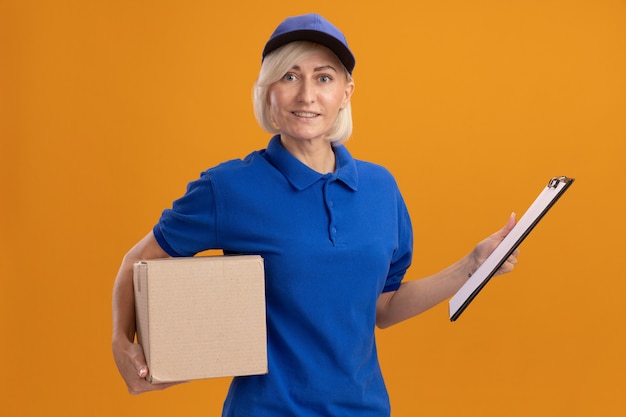 Glimlachende blonde bezorger van middelbare leeftijd in blauw uniform en pet met kartonnen doos en klembord