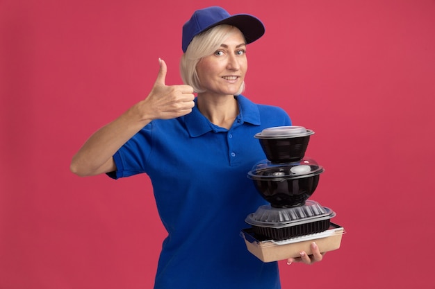 Glimlachende blonde bezorger van middelbare leeftijd in blauw uniform en pet met papieren voedselpakket en voedselcontainers met duim omhoog geïsoleerd op roze muur