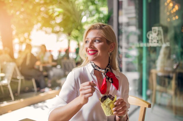 Glimlachende blonde jonge vrouw die chocolademuffin in document houder eet