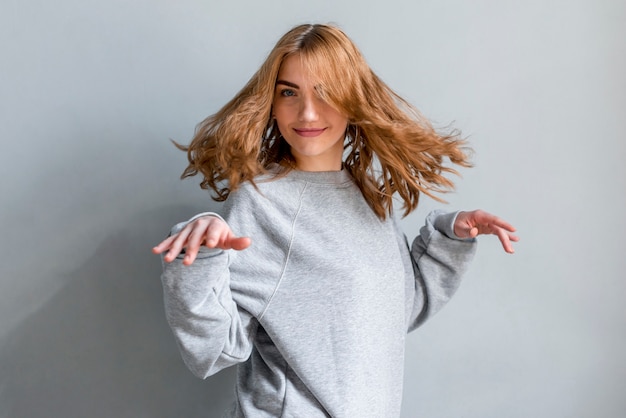 Gratis foto glimlachende blonde jonge vrouw die tegen grijze muur dansen