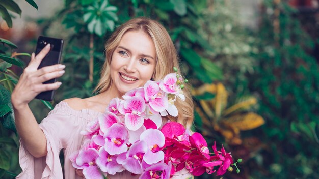 Glimlachende blonde jonge vrouw die verse orchidee houdt die in hand zelfportret van mobiele telefoon neemt