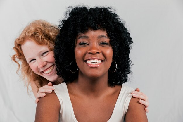 Glimlachende blonde jonge vrouw die zich achter de Afrikaanse vriend tegen grijze achtergrond bevindt