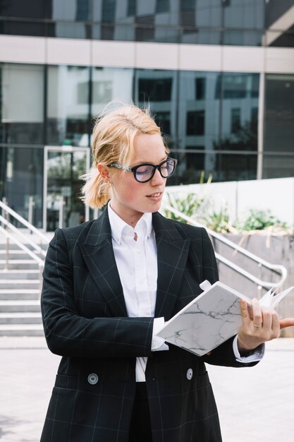 Glimlachende blonde jonge vrouw die zich buiten het gebouw bevindt dat het agenda controleert