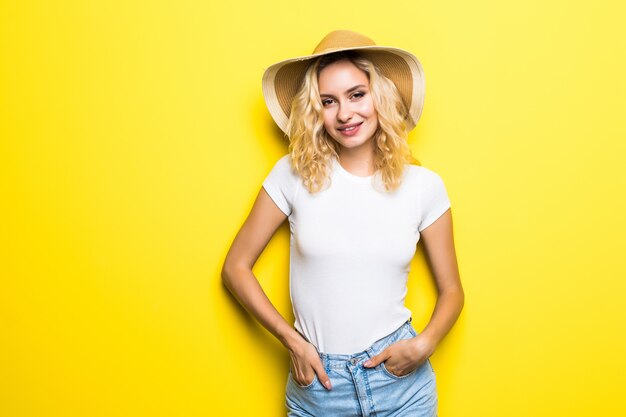 Glimlachende blonde jonge vrouw in strohoed, jeansborrels, wit overhemd op gele muur.
