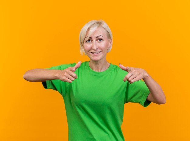 Glimlachende blonde Slavische vrouw die op middelbare leeftijd voorzijde bekijkt die u gebaar doet dat op gele muur wordt geïsoleerd