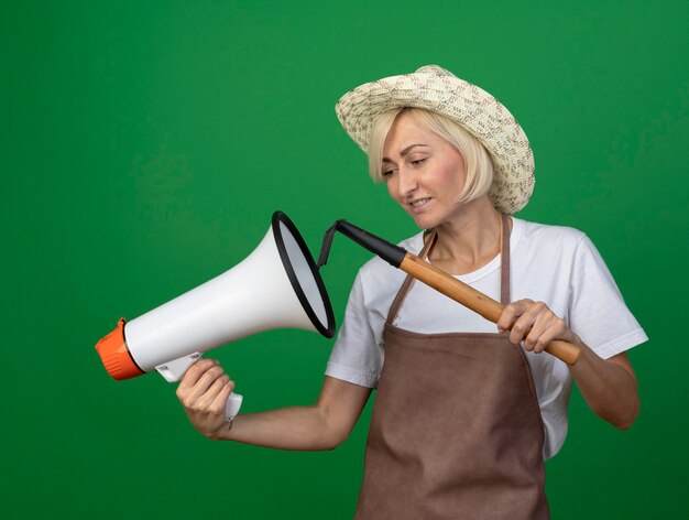 Glimlachende blonde tuinman vrouw van middelbare leeftijd in uniform dragen hoed houden en kijken naar spreker houden hark in de buurt van het geïsoleerd op groene muur