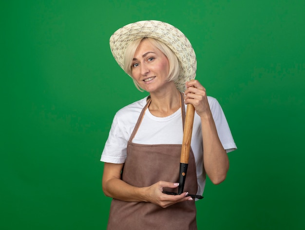 Glimlachende blonde tuinmanvrouw van middelbare leeftijd in uniform met hoed met hark ondersteboven geïsoleerd op groene muur met kopieerruimte