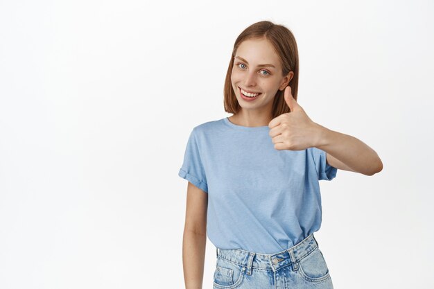 Glimlachende blonde vrouw die duim opsteekt en goedkeurend knikt, je prijst, een compliment maakt, iets goeds aanbeveelt, tegen een witte muur staat