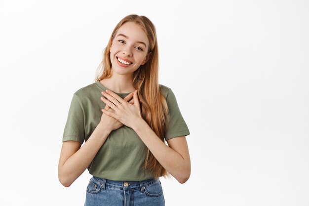 Glimlachende blonde vrouw die handen op het hart houdt en er aangeraakt en gevleid uitziet, zeg dankjewel, blij en opgetogen, staande tegen een witte muur