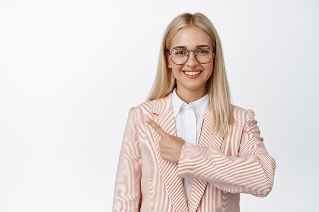 Glimlachende blonde vrouw met een bril die met de vinger naar links wijst, een bril en een roze pak op wit draagt.