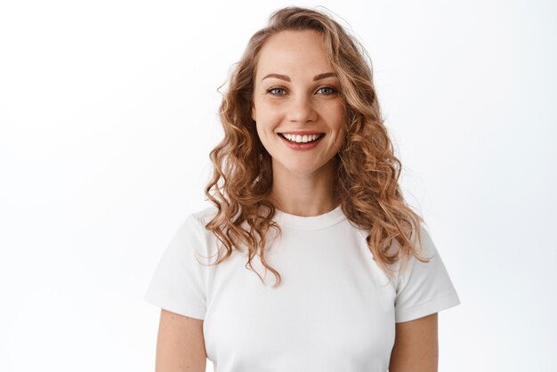 Glimlachende blonde vrouw met witte perfecte glimlach en natuurlijk gezicht die gelukkig en zelfverzekerd kijkt naar de camera die in een t-shirt staat tegen een witte achtergrond