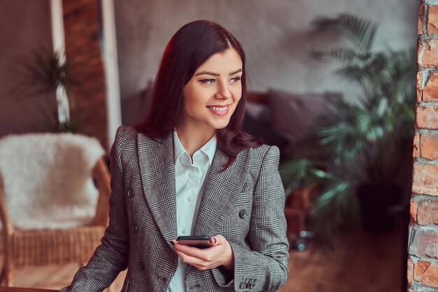 Glimlachende charmante brunette vrouw gekleed in een grijze elegante jas met smartphone in een kamer met loft interieur, wegkijkend.