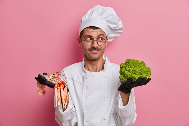 Glimlachende chef-kok kijkt graag opzij, draagt een koksmuts en uniform, houdt groene broccoli en kreeft vast, stelt het beste menu voor vegetariërs in het café voor