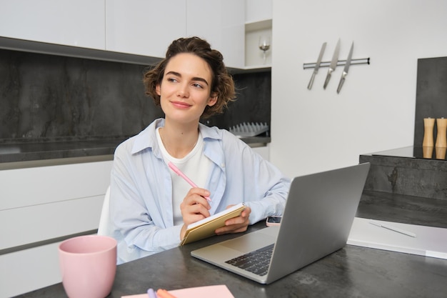 Glimlachende creatieve vrouw met pen en notitieboek ziet er bedachtzaam uit werkt thuis met laptop in de keuken