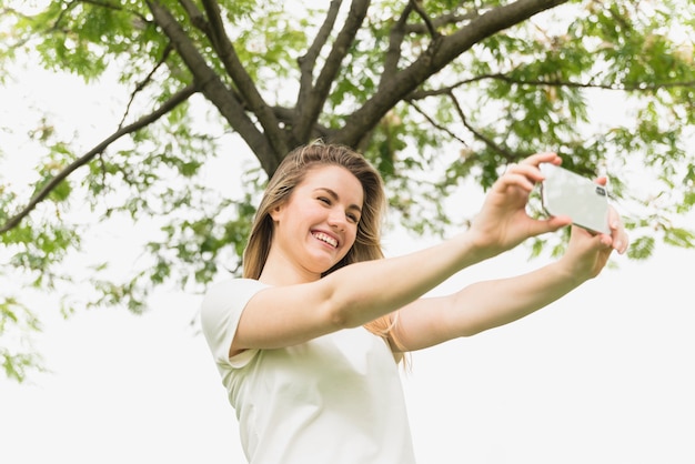 Glimlachende dame die selfie op mobiele telefoon dichtbij boom nemen