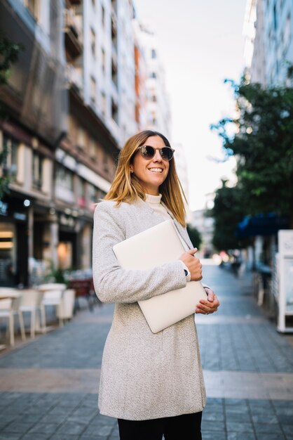 Glimlachende elegante jonge vrouw met zonnebril en laptop op straat in stad