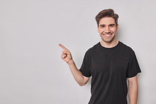 Glimlachende Europese man met blije uitdrukking wijst op lege kopieerruimte toont promotieaanbieding aantoont dat advertentie casual zwart t-shirt draagt dat op witte achtergrond wordt geïsoleerd. Kijk hiernaar.