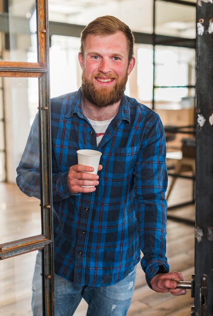 Glimlachende gebaarde mens die beschikbare koffiekop houden terwijl het openen van deur