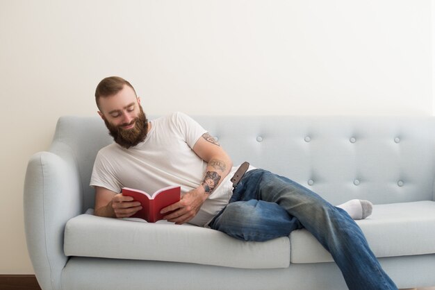 Glimlachende gebaarde mens die op bank ligt en boek leest